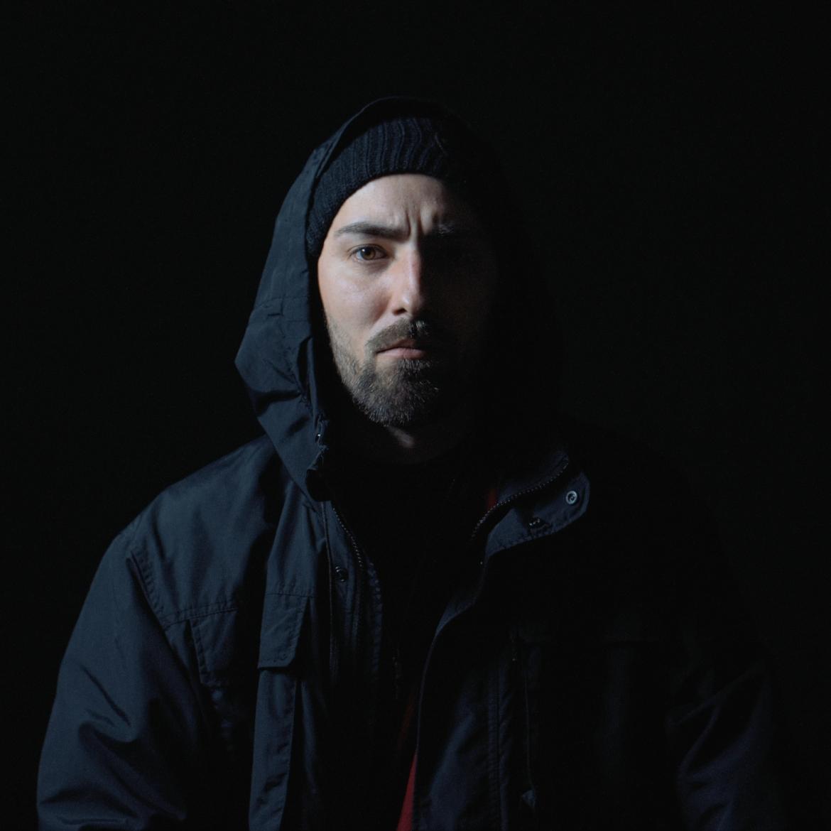 Bild des Künstlers "Accid1" in schwarzer Kapuzenjacke vor schwarzem Hintergrund.